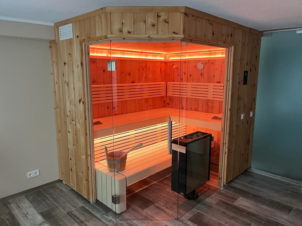 Eck-Sauna aus hellem Fichtenholz mit Glastür, warmes Rotlicht, Kelo-Ofen